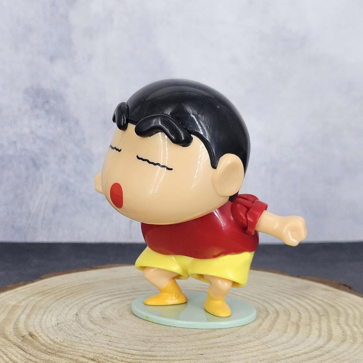 Toy figure of a character with black hair, red shirt, and yellow shorts on a wooden surface with a gray background.